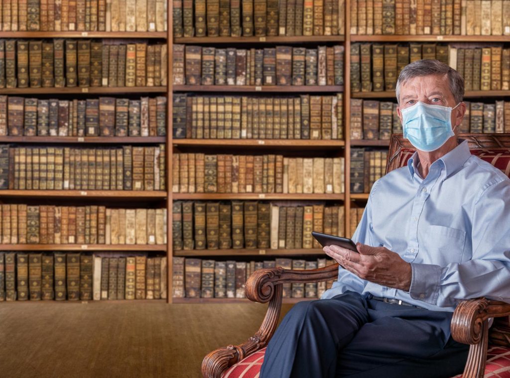 Stock photo of Senior retired man reading an eBook in his antique library of old books with a face mask against coronavirus